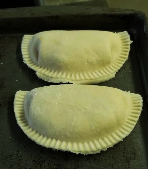 Two uncooked empanadas on a baking tray, shaped with a crimped edge and ready for baking.