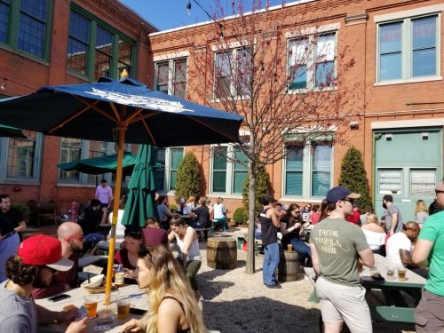 A lively outdoor patio scene with people dining and socializing under umbrellas, surrounded by brick buildings and trees.