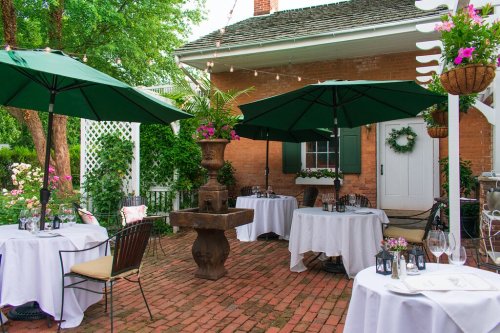 Outdoor dining area with green umbrellas, white tablecloths, and potted plants, set against a charming brick backdrop.