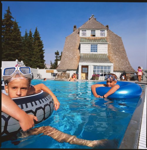 Two children in inflatable rings relax in a pool, with a large building and trees in the background on a sunny day.