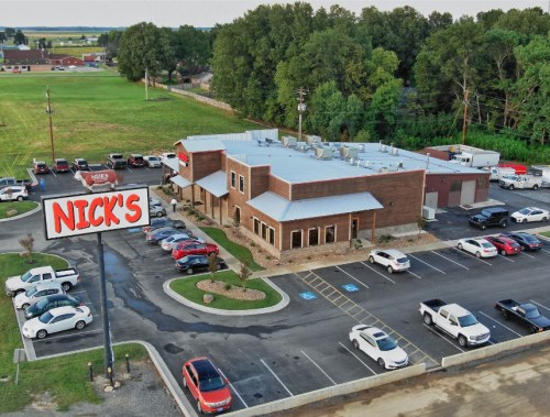 Aerial view of Nick's restaurant with a large sign, surrounded by a parking lot filled with cars and green fields in the background.