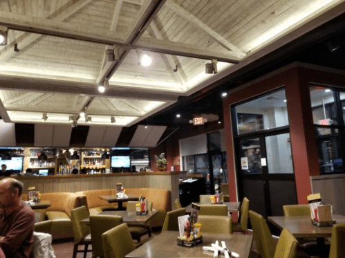 Interior of a restaurant featuring booths, tables, and a bar area, with warm lighting and a wooden ceiling.