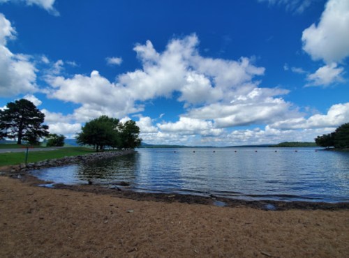 A serene lakeside view with sandy shore, trees, and a blue sky filled with fluffy clouds.