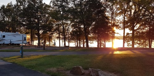 A serene campsite at sunset, featuring an RV surrounded by trees and a view of the water in the background.