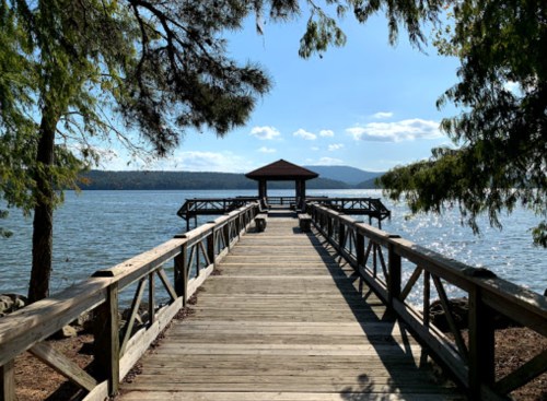 A wooden dock extends into a calm lake, surrounded by trees and mountains under a clear blue sky.