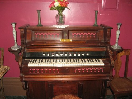 A vintage wooden organ with decorative details, candles, and a vase of flowers against a pink wall.