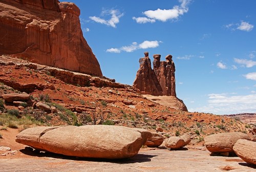 Three tall rock formations rise against a blue sky, surrounded by red desert landscape and scattered boulders.