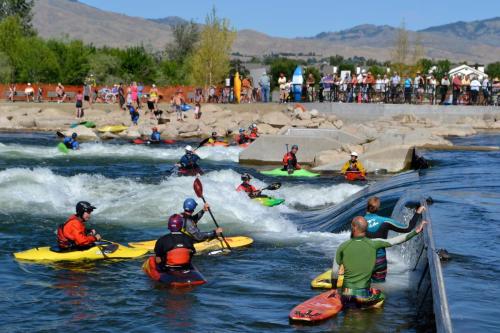 A lively scene of kayakers navigating rapids, with spectators watching from the shore in a sunny outdoor setting.