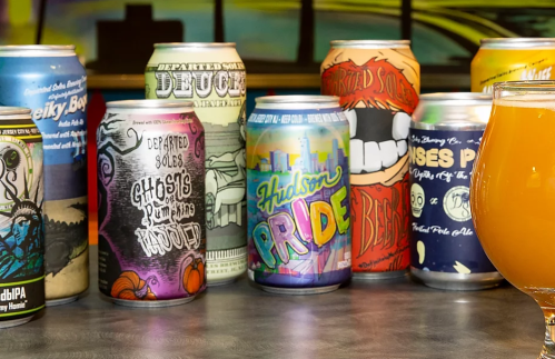 A variety of colorful beer cans displayed on a table, with a glass of beer in the foreground.