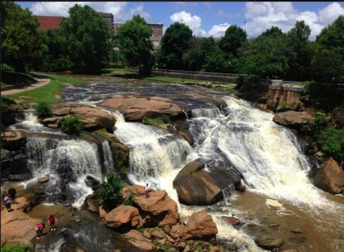 A scenic view of a waterfall cascading over rocks, surrounded by lush greenery and a clear blue sky.