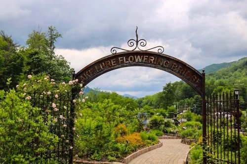 A wrought iron archway reads "Nature Flowering Bridge," surrounded by lush greenery and blooming flowers under a cloudy sky.