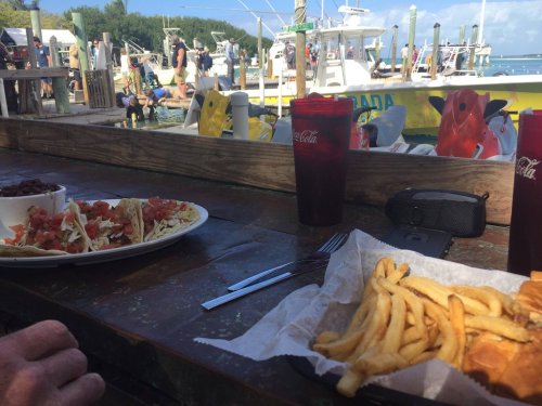 A table with a plate of tacos, fries, and drinks, overlooking a busy marina with boats and people.