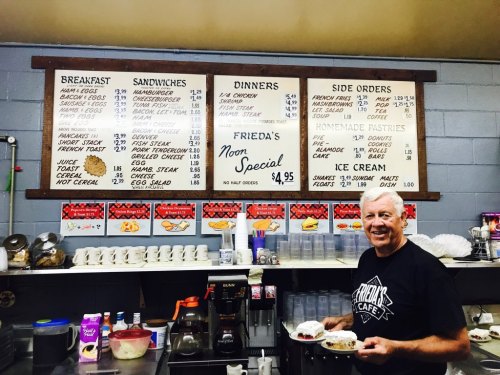A smiling man holds two plates of food in a café with a menu board displaying various meal options and prices.