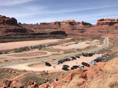 A scenic view of a desert landscape with a river, parking area, and several vehicles near red rock formations.