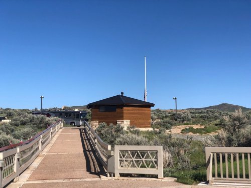 A wooden building with a flagpole, surrounded by greenery and a clear blue sky, accessible via a bridge.