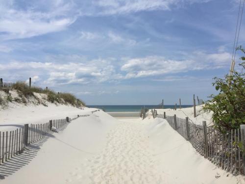 A sandy path leads to a calm beach under a blue sky with scattered clouds and gentle dunes on either side.
