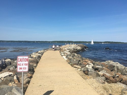 A rocky jetty extends into the water, with a caution sign and people walking along the path under a clear blue sky.