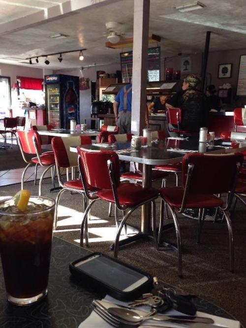A cozy diner interior with red chairs, tables, and a drink on the table, featuring patrons in the background.