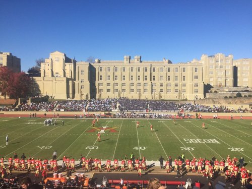 A football game in progress with players on the field and a large crowd in the stands, set against a historic building backdrop.