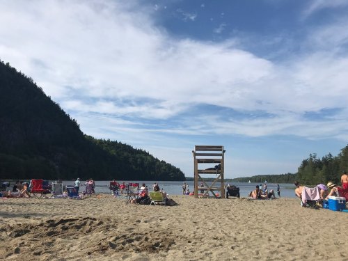 A sandy beach with people relaxing, a lifeguard tower, and a scenic backdrop of trees and mountains under a blue sky.
