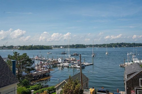 A serene waterfront view with boats docked, lush greenery, and a clear blue sky with scattered clouds.
