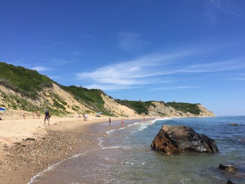A sunny beach scene with people walking along the shore, cliffs in the background, and gentle waves lapping at the sand.