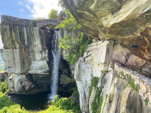 A waterfall cascades down rocky cliffs surrounded by lush greenery and a stone pathway under a blue sky.