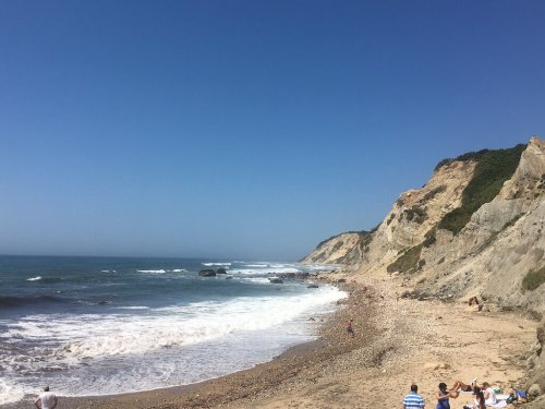A sunny beach scene with waves crashing on the shore, rocky cliffs in the background, and people enjoying the sand.