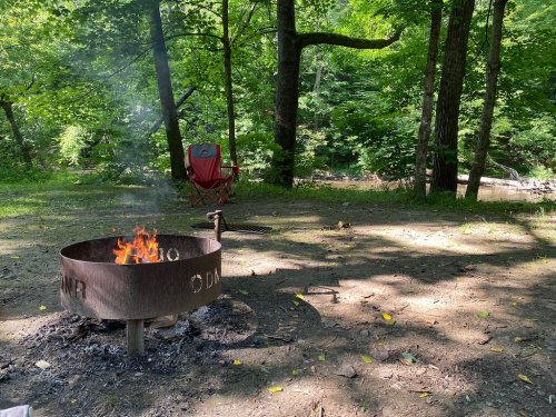 A campfire burns in a metal ring surrounded by trees, with a red chair nearby and a stream in the background.
