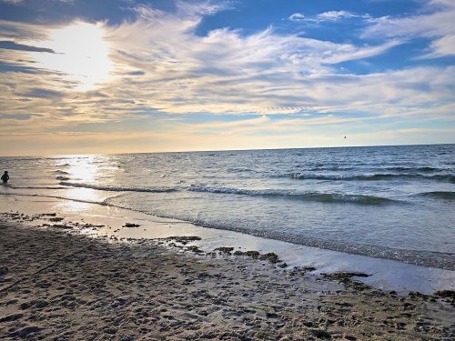 A serene beach scene at sunset, with gentle waves lapping at the shore and a person walking along the water's edge.