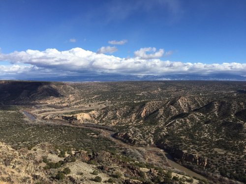 A panoramic view of a rugged landscape with rolling hills, a winding river, and a blue sky dotted with clouds.