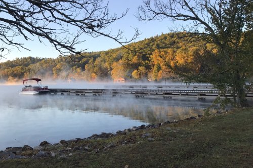 A serene lake scene at dawn, with mist rising over the water and a boat docked by a tree-lined shore.