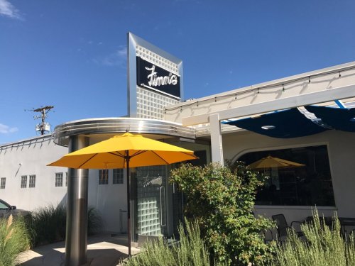 Exterior of a restaurant with a bright yellow umbrella, modern signage, and a clear blue sky in the background.