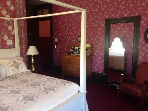 A vintage bedroom with a four-poster bed, floral wallpaper, a mirror, and a wooden dresser.