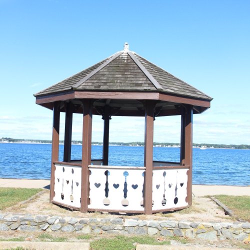 A wooden gazebo with a shingled roof by a calm lake under a clear blue sky.