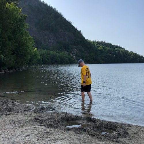 A person in a yellow shirt wades into a calm lake, surrounded by trees and a rocky hillside.