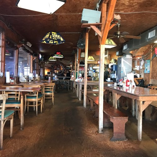 Interior of a rustic restaurant with wooden tables, chairs, and vintage decor, featuring a bar and hanging lights.