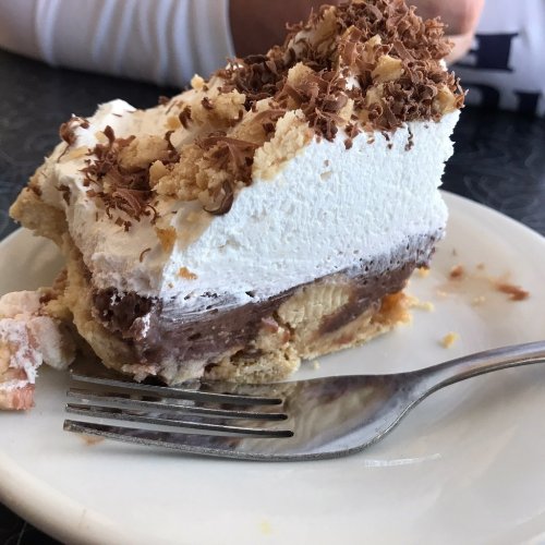 A slice of layered dessert with chocolate, cream, and chocolate shavings on top, served on a white plate with a fork.