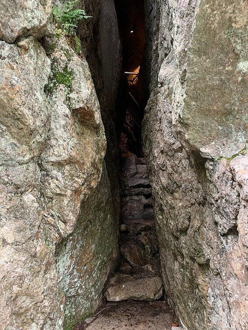 Narrow stone passageway between large rocks, leading to a dimly lit area with steps.