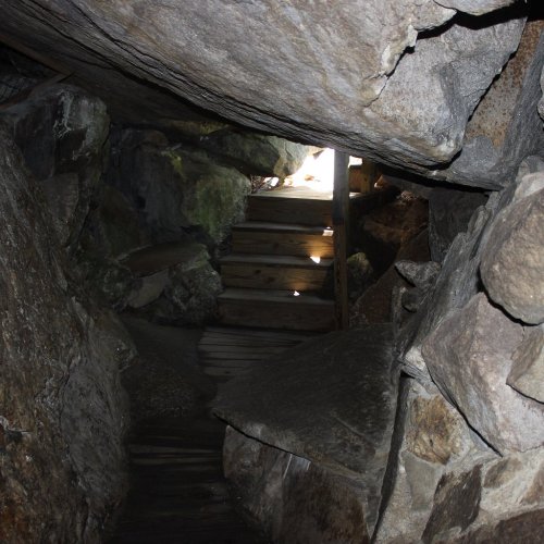 A narrow passageway between large rocks, leading to a wooden staircase illuminated by light at the end.