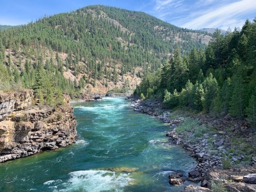 A serene river flows through a lush green landscape, surrounded by mountains and trees under a clear blue sky.