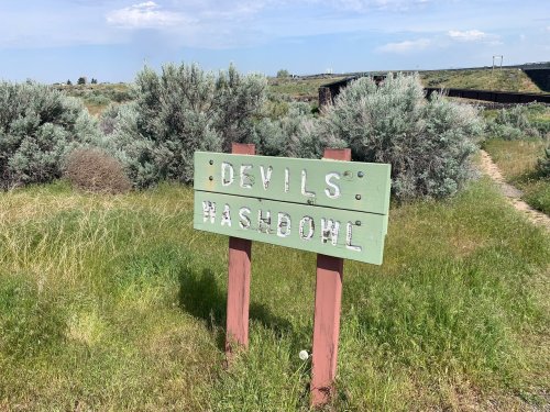 Sign reading "Devils Washbowl" in a grassy area with shrubs and a blue sky in the background.