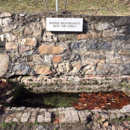 Stone structure with a sign reading "George Washington's Bath Tub (1748)" and water with fallen leaves inside.