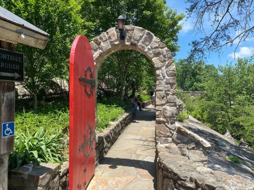 A red door in a stone archway leads to a scenic pathway surrounded by greenery and trees on a sunny day.