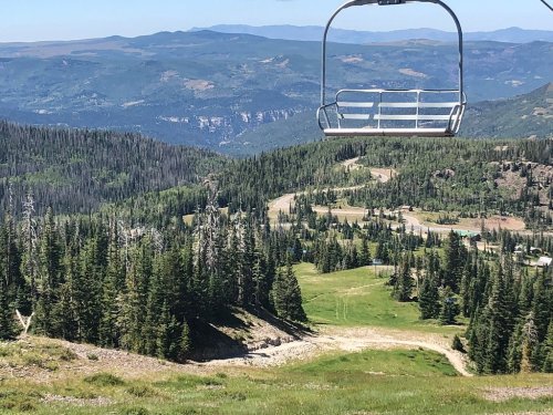 A scenic view from a ski lift overlooking green hills, trees, and distant mountains under a clear blue sky.