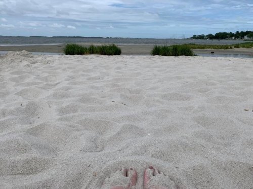 Feet resting in soft sand with a view of a calm beach and water under a cloudy sky.
