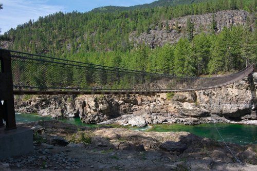 A suspension bridge spans a river, surrounded by lush green trees and rocky cliffs under a clear blue sky.