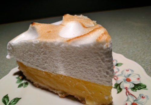 A slice of lemon meringue pie on a floral plate, featuring a golden filling and fluffy white meringue topping.