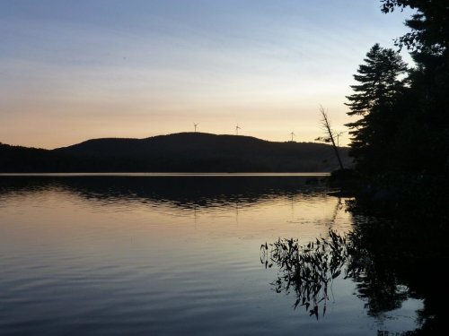 A serene lake at sunset, reflecting the sky and surrounded by hills and trees, with wind turbines in the distance.