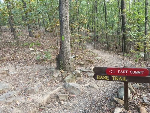A trail sign points to "East Summit" and "Base Trail" in a wooded area with trees and rocky terrain.